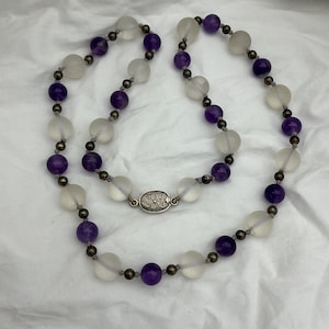 May include: A necklace with alternating frosted white and deep purple beads. The beads are strung with small, dark grey beads. The necklace is fastened with a silver-coloured clasp with an ornate design. The jewellery is displayed on a white surface.