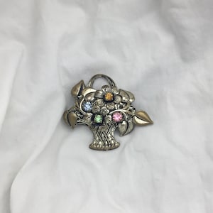 May include: A silver-toned brooch shaped like a basket overflowing with colorful flowers. The flowers are made of rhinestones in shades of pink, blue, green, and orange.