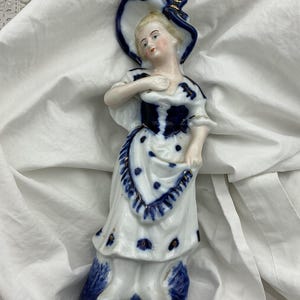 Op de afbeelding: Een porseleinen beeldje van een persoon met een hoed met brede rand, een lijfje en een rok met een blauw-wit bloemmotief. Het figuur heeft goudkleurige accenten op de hoed, het lijfje en de zoom van de rok en staat op een blauwe voet.