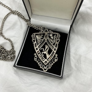 May include: A silver-toned pendant necklace with a shield-shaped design, featuring a horse, thistle, and other heraldic elements. The necklace is displayed in a black box with a white interior. The chain is visible to the left.