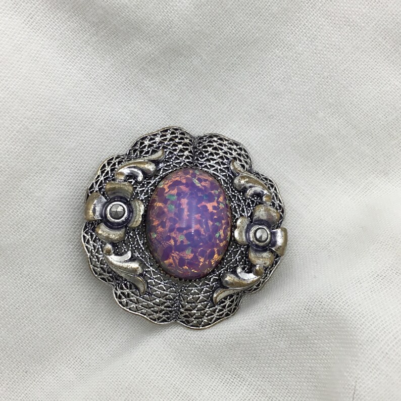 Puede incluir: Un broche de plata con un cabuj&oacute;n de &oacute;palo iridiscente de forma ovalada engastado en un dise&ntilde;o de filigrana. El broche presenta dos peque&ntilde;os detalles florales a cada lado del &oacute;palo.