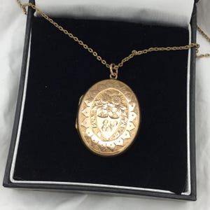 Può includere: Una collana con un ciondolo in oro, decorato con un motivo floreale e le iniziali "Eva" incise sul davanti. Il ciondolo è in una scatola di velluto nero.