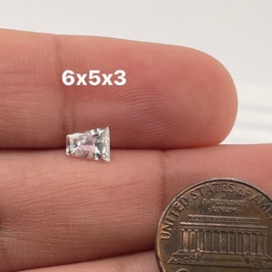 Peut inclure: Une pierre précieuse rectangulaire transparente avec une légère teinte rose, mesurant 6x5x3 millimètres, est montrée sur un doigt à côté d'un penny américain.