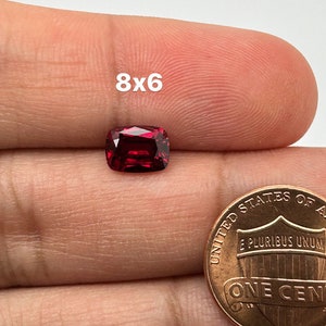 May include: A deep red gemstone with a cushion cut, measuring 8x6 millimeters, is held in a hand next to a US penny for size comparison.
