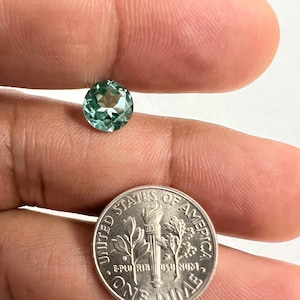 Puede incluir: Una piedra preciosa redonda y facetada de color verde se sostiene en una mano, junto a una moneda de diez céntimos de euro para comparar el tamaño.