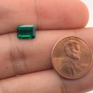 May include: A vibrant green emerald gemstone, cut in a rectangular shape, is shown next to a US penny for size comparison.
