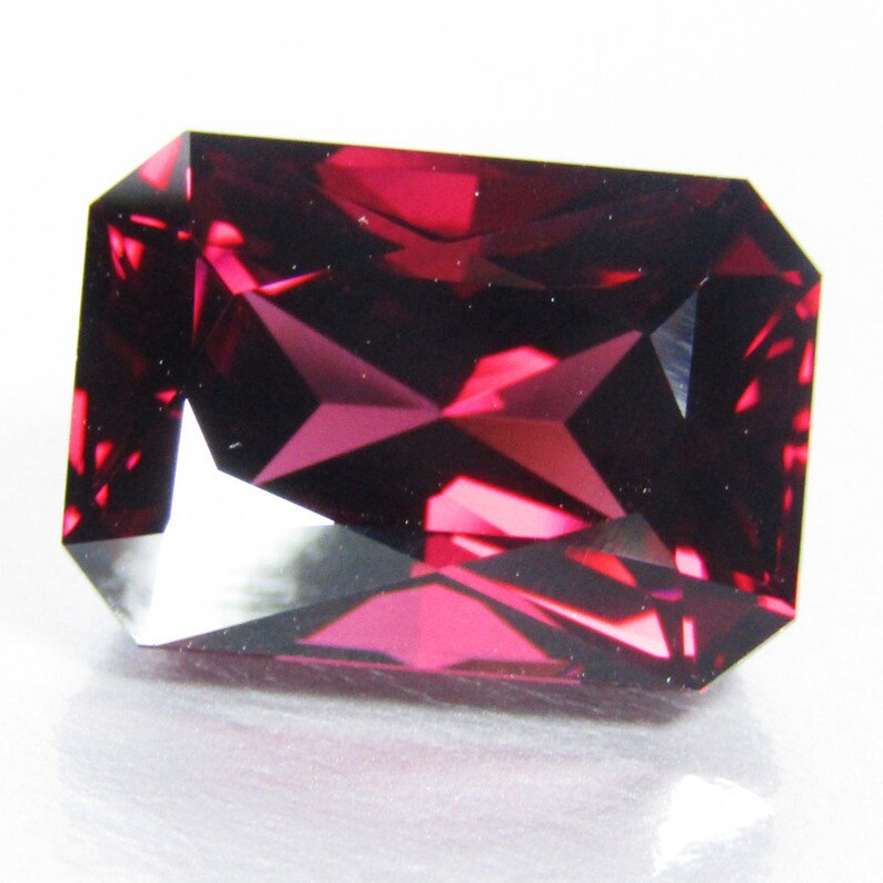 Peut inclure: Une pierre pr&eacute;cieuse rouge fonc&eacute; de forme rectangulaire et &agrave; facettes. La pierre est pos&eacute;e sur un fond blanc.