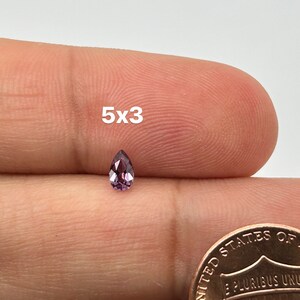 May include: A pear-shaped gemstone with a purple hue, measuring 5x3 millimeters, is shown on a finger.