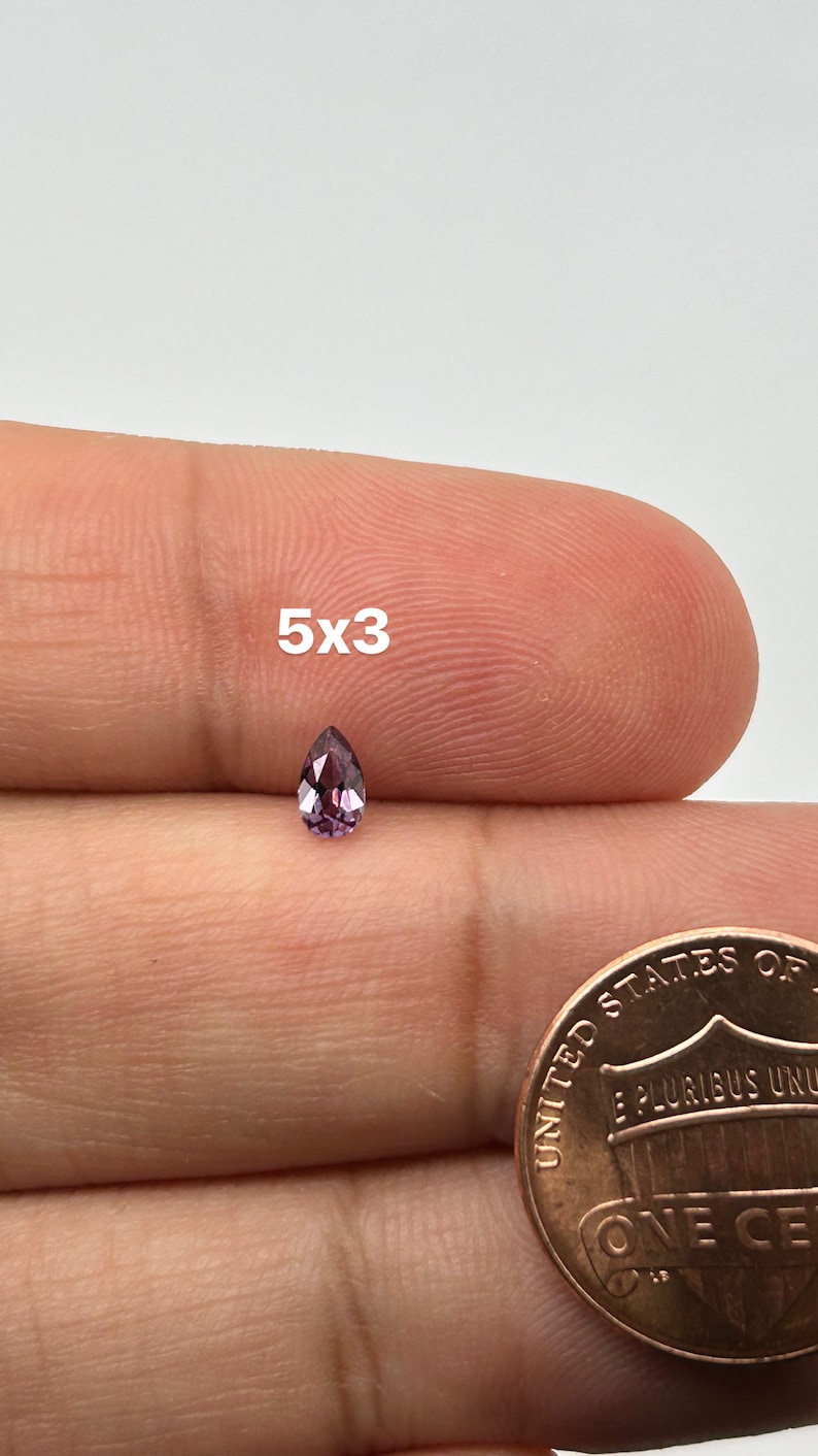 May include: A pear-shaped gemstone with a purple hue, measuring 5x3 millimeters, is shown on a finger.