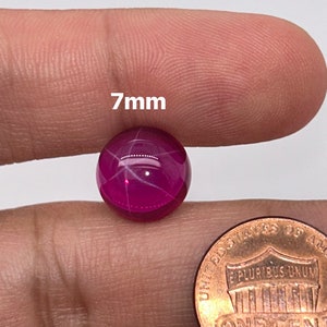 May include: A 7mm round, pink ruby cabochon gemstone with a star-like pattern in the center. The stone is held in a person's hand for size comparison.
