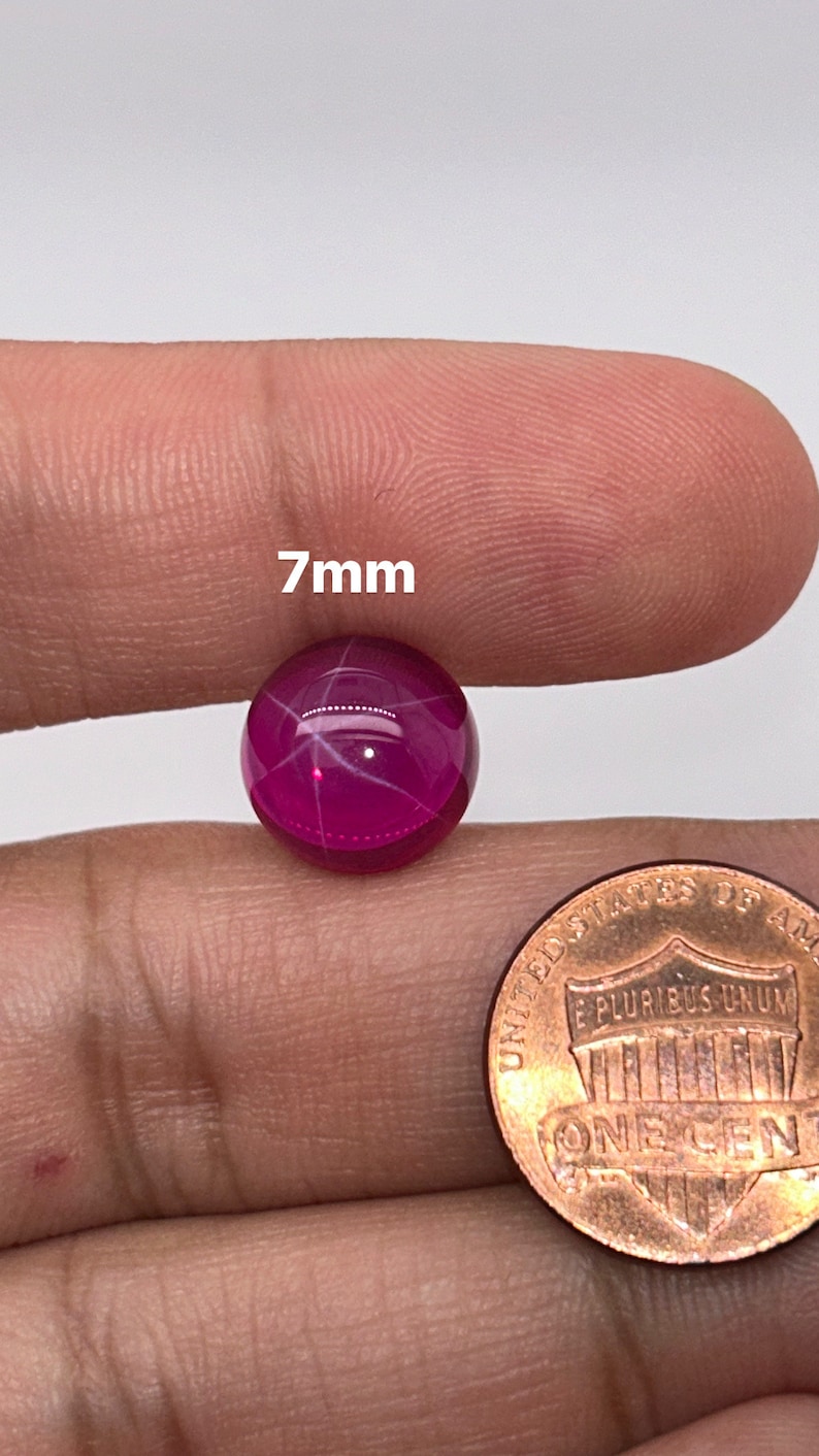 May include: A 7mm round, pink ruby cabochon gemstone with a star-like pattern in the center. The stone is held in a person's hand for size comparison.