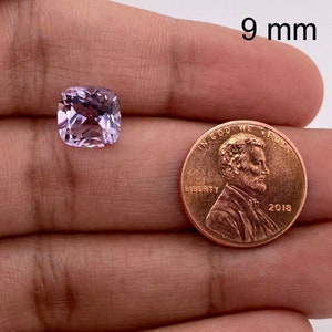 May include: A light purple cushion-cut gemstone measuring 9 mm in diameter, held in a hand next to a US penny for size comparison.