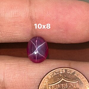 May include: A pink star ruby gemstone with a six-pointed star in the center. The gemstone is approximately 10x8 mm in size and is being held in a hand next to a US penny for size comparison.