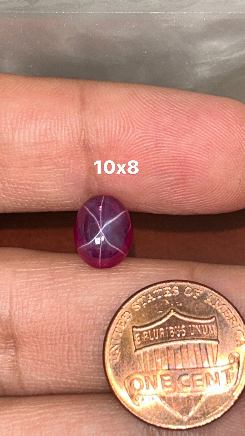 May include: A pink star ruby gemstone with a six-pointed star in the center. The gemstone is approximately 10x8 mm in size and is being held in a hand next to a US penny for size comparison.