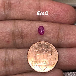 May include: A pink star ruby gemstone measuring 6x4 millimeters. The gemstone is held in a hand for size comparison next to a US penny.