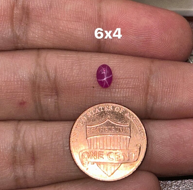 May include: A pink star ruby gemstone measuring 6x4 millimeters. The gemstone is held in a hand for size comparison next to a US penny.
