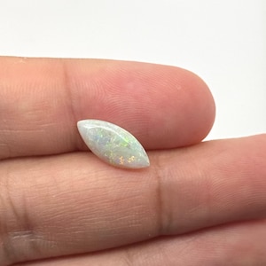 May include: A white opal gemstone with a teardrop shape. The opal displays iridescent flashes of green, blue, and gold across its surface. The gemstone is held in a hand, showcasing its size and color variations.