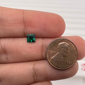 May include: A vibrant green square gemstone, approximately the size of a UK penny, is held in a hand. The gemstone is faceted and has a brilliant shine.