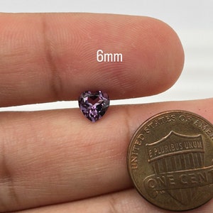 K&ouml;nnte beinhalten: Ein 6 mm gro&szlig;er herzf&ouml;rmiger violetter Edelstein. Der Edelstein wird von einer Hand gehalten, und ein US-Cent befindet sich im Hintergrund zum Gr&ouml;&szlig;envergleich.