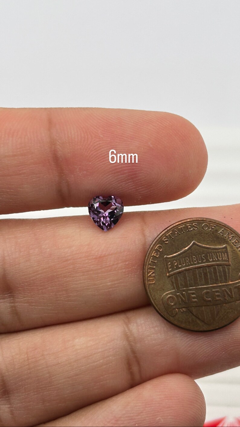 K&ouml;nnte beinhalten: Ein 6 mm gro&szlig;er herzf&ouml;rmiger violetter Edelstein. Der Edelstein wird von einer Hand gehalten, und ein US-Cent befindet sich im Hintergrund zum Gr&ouml;&szlig;envergleich.