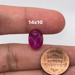 May include: A pink star ruby cabochon gemstone with a 14x10 mm size, shown next to a US penny for scale.