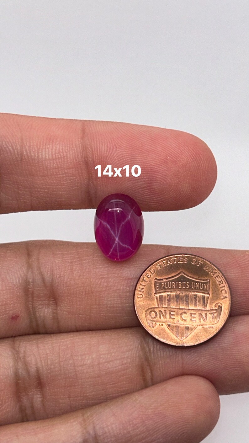 May include: A pink star ruby cabochon gemstone with a 14x10 mm size, shown next to a US penny for scale.