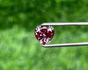 Può includere: Una gemma a forma di cuore tenuta da una pinzetta. La gemma è di un colore bordeaux intenso con macchie iridescenti blu e bianche. Lo sfondo è un verde sfocato.