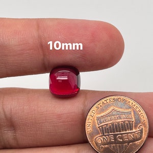 May include: A deep red, 10mm cabochon gemstone, shown next to a US penny for size comparison.