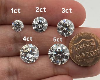May include: Five round-cut diamonds, labelled 1ct to 5ct, are shown next to a penny for scale. The diamonds are clear and faceted, reflecting light. The image is taken against a neutral background. The penny is a British one penny coin.