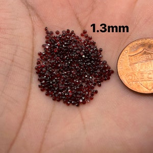 May include: A pile of small, dark red, faceted gemstones, approximately 1.3 mm in size, next to a US penny for scale.