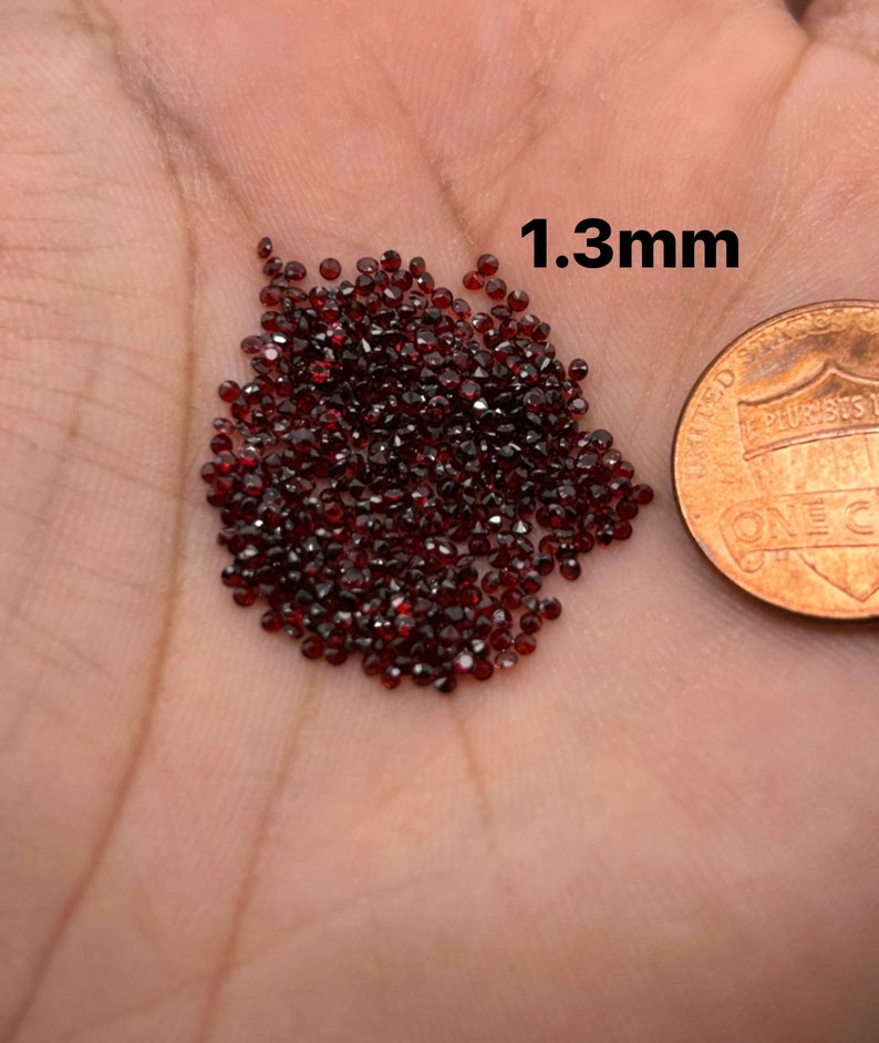 May include: A pile of small, dark red, faceted gemstones, approximately 1.3 mm in size, next to a US penny for scale.