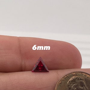 May include: A 6mm triangular red gemstone, held in a hand for size comparison, next to a US penny.