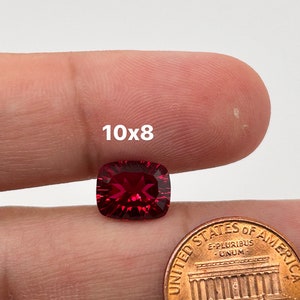 May include: A deep red gemstone with a cushion cut, measuring 10x8 millimeters, is held in a hand next to a US penny for size comparison.