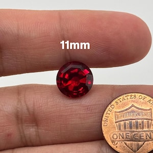 May include: A deep red gemstone, approximately 11mm in diameter, is shown on a finger. The gemstone is faceted and has a round shape. A UK penny is shown for size comparison.