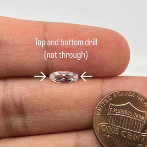 May include: A clear, faceted gemstone with a slight pink hue. The gemstone is held in a hand next to a US penny for size comparison. The gemstone has drill holes at the top and bottom, but the holes do not go all the way through.