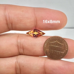 May include: A faceted, kite-shaped gemstone, measuring 16x8mm, with a blend of orange, yellow, and pink hues. A US one-cent coin is placed next to the gemstone for size comparison. The gemstone is held between two fingers.