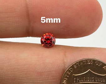 Puede incluir: Una piedra preciosa roja redonda de 5 mm con facetas, sostenida en una mano para comparar el tamaño junto a un centavo de dólar estadounidense.