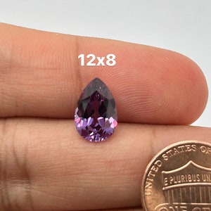May include: A pear-shaped purple gemstone measuring 12x8 mm. The gemstone is held in a hand with a US penny for scale.