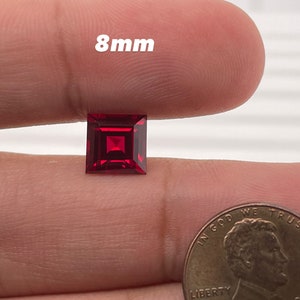 May include: A deep red square-shaped gemstone measuring 8mm in size. The gemstone is held in a hand for size comparison.