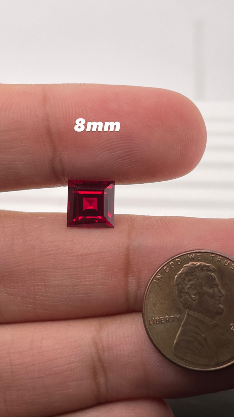 May include: A deep red square-shaped gemstone measuring 8mm in size. The gemstone is held in a hand for size comparison.