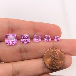 May include: Five pink cushion-cut gemstones of varying sizes, displayed on a hand with a US penny for scale.  "In God We Trust" and "Liberty" are visible on the coin.