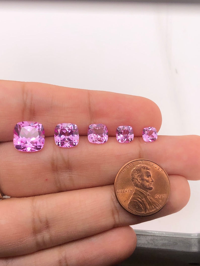 May include: Five pink cushion-cut gemstones of varying sizes, displayed on a hand with a US penny for scale.  "In God We Trust" and "Liberty" are visible on the coin.