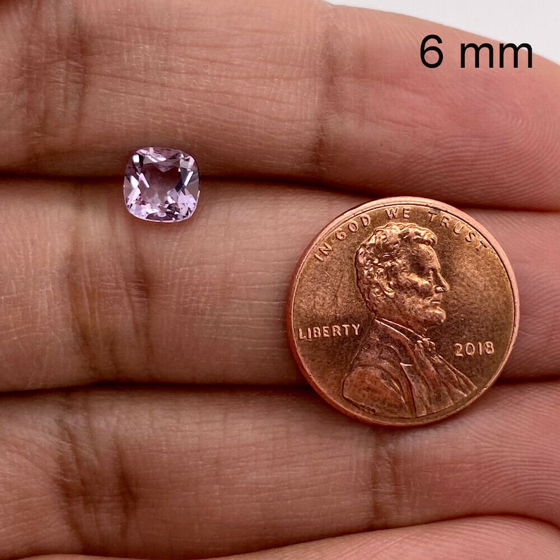 May include: A light pink cushion-cut gemstone measuring 6 mm in diameter, shown next to a US penny for size comparison.