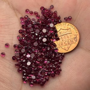 May include: A collection of small, faceted, deep red gemstones, possibly garnets, are scattered across a hand. A U.S. one-cent coin is placed among the gems for size comparison. The gemstones have a brilliant, reflective quality.