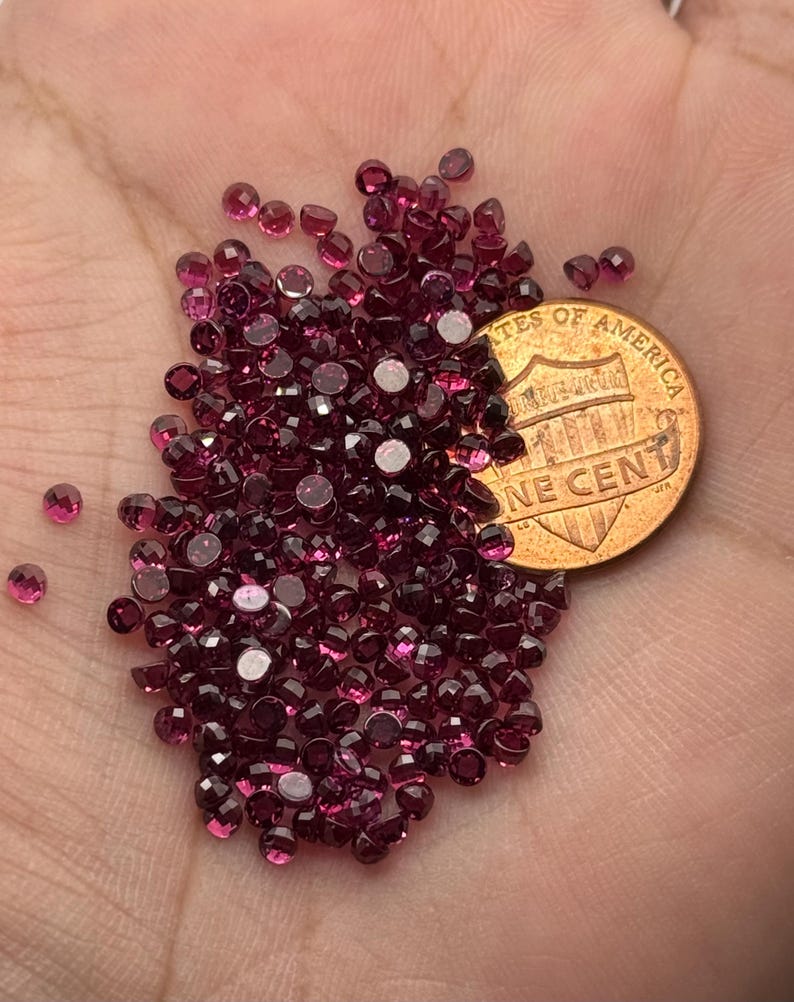 May include: A collection of small, faceted, deep red gemstones, possibly garnets, are scattered across a hand. A U.S. one-cent coin is placed among the gems for size comparison. The gemstones have a brilliant, reflective quality.