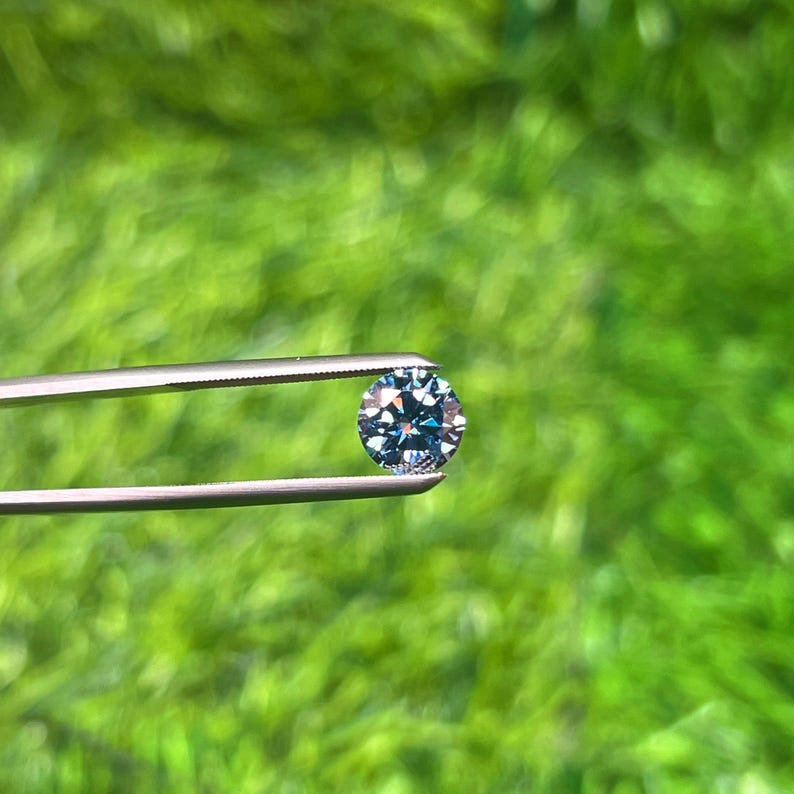 Op de afbeelding: Een ronde, gefacetteerde blauwe edelsteen wordt vastgehouden door een zilveren pincet tegen een wazige groene achtergrond. De edelsteen heeft meerdere facetten die licht reflecteren, waardoor een sprankelend effect ontstaat. De pincet is scherp.
