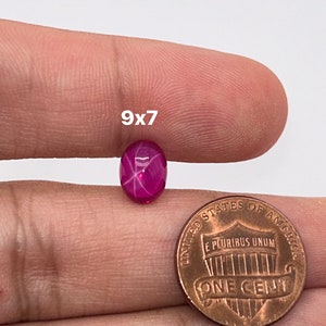 May include: A 9x7mm pink star ruby gemstone with a star-like pattern in the center. The gemstone is being held in a hand next to a US penny for size comparison.