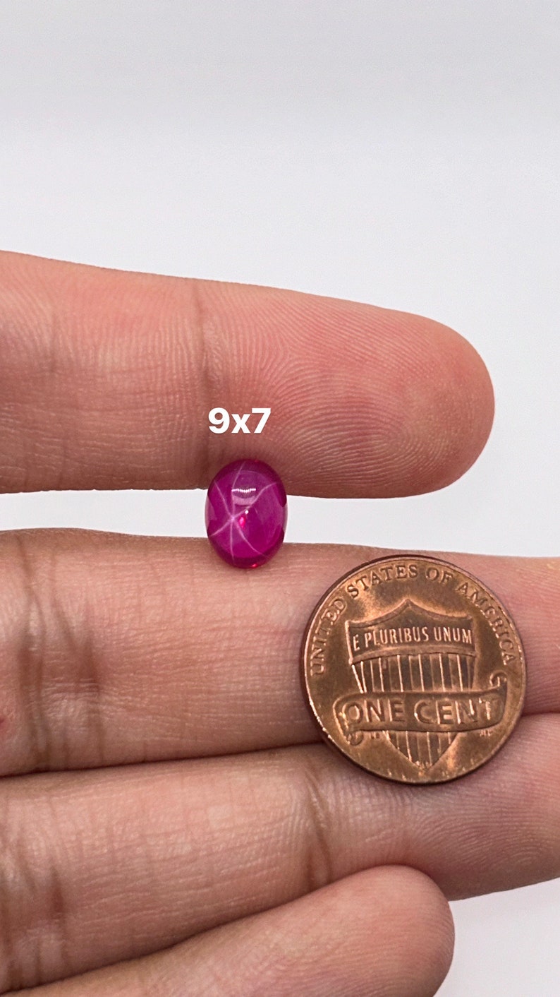 May include: A 9x7mm pink star ruby gemstone with a star-like pattern in the center. The gemstone is being held in a hand next to a US penny for size comparison.
