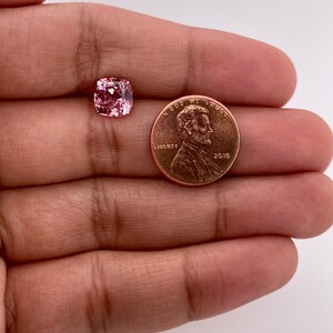May include: A pink gemstone with a cushion cut, next to a US penny for size comparison.
