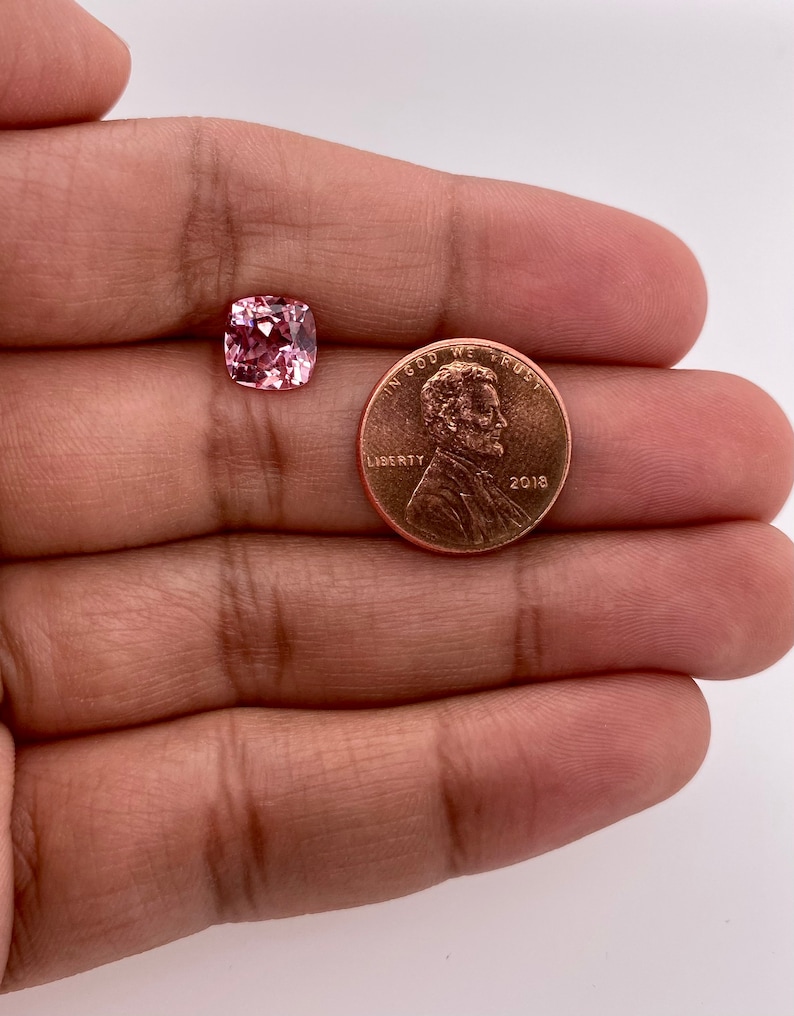 May include: A pink gemstone with a cushion cut, next to a US penny for size comparison.